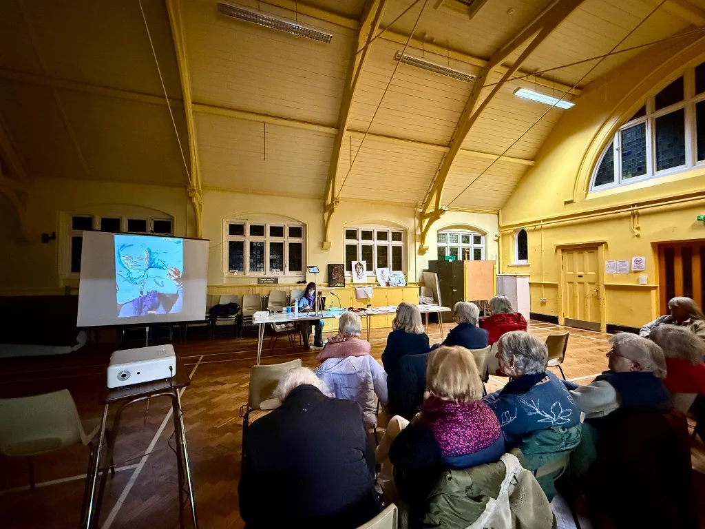 An audience seated in a hall watching an artist at a table, with a portrait drawing projected onto a screen beside them