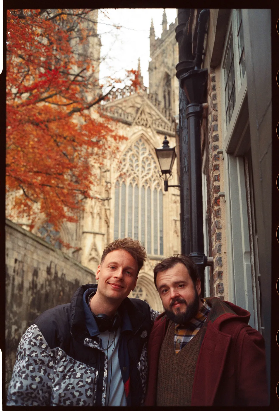 Two men in coats stand together in a narrow lane beside a cathedral, with a tree in autumn colours behind them