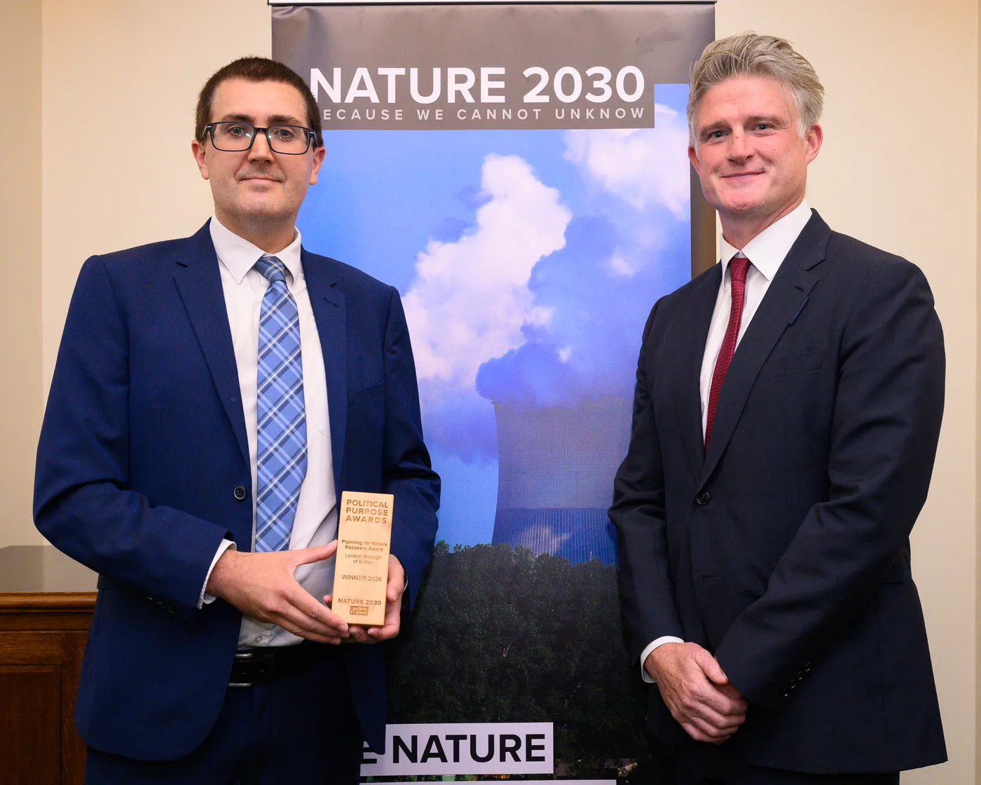 Two men in suits stand either side of the wooden Political Purpose Awards trophy in front of a Nature 2030 banner